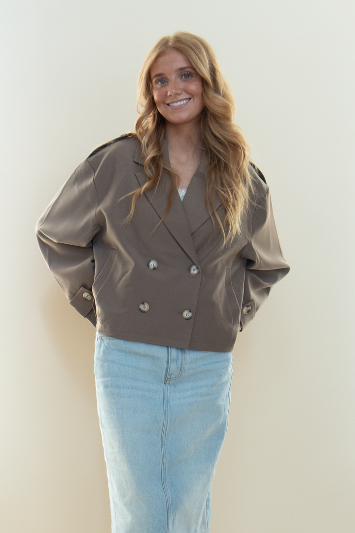 Close up of taupe cropped double breasted trench jacket with notch lapel and button detail, worn over a light wash denim skirt