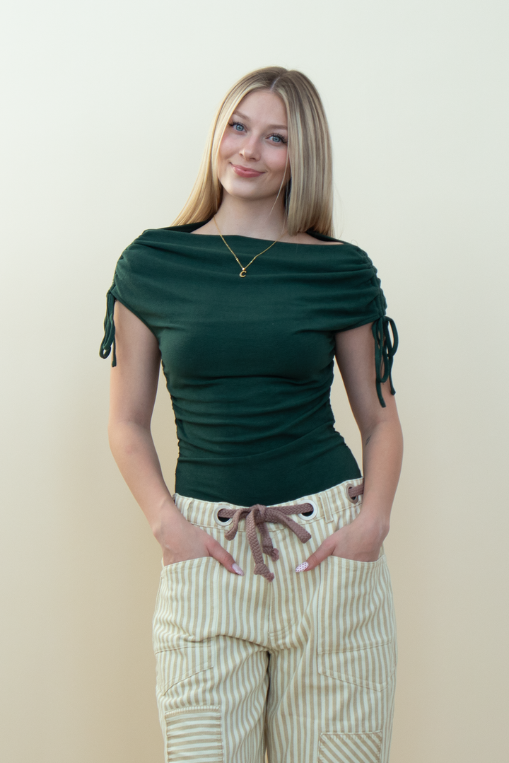 A woman modeling a dark green off-the-shoulder top with side ties and cream and tan vertically striped pants, standing against a plain beige background.
