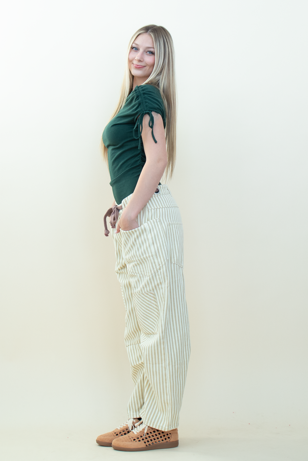 A woman posing in a studio setting, wearing a dark green short-sleeve top, cream and white vertically striped cargo pants, and brown crocheted sneakers.
