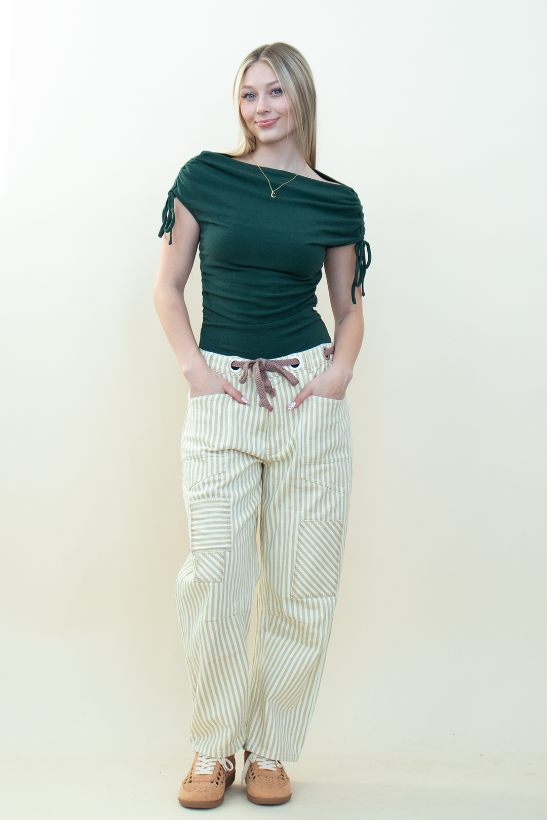 A woman modeling a dark green boatneck bodysuit and cream and white vertically striped drawstring pants, paired with brown shoes, against a plain cream background.