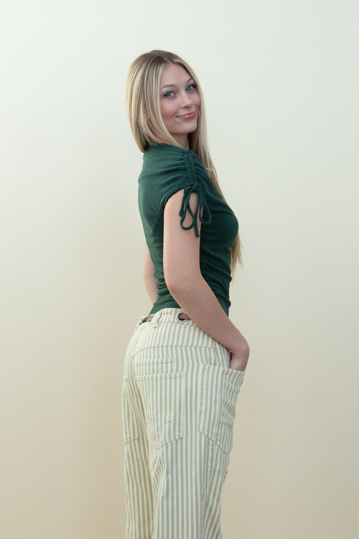 A woman with blonde hair is shown from the side, wearing a dark green short-sleeve top with side ties and cream-colored striped pants, standing against a plain beige background.