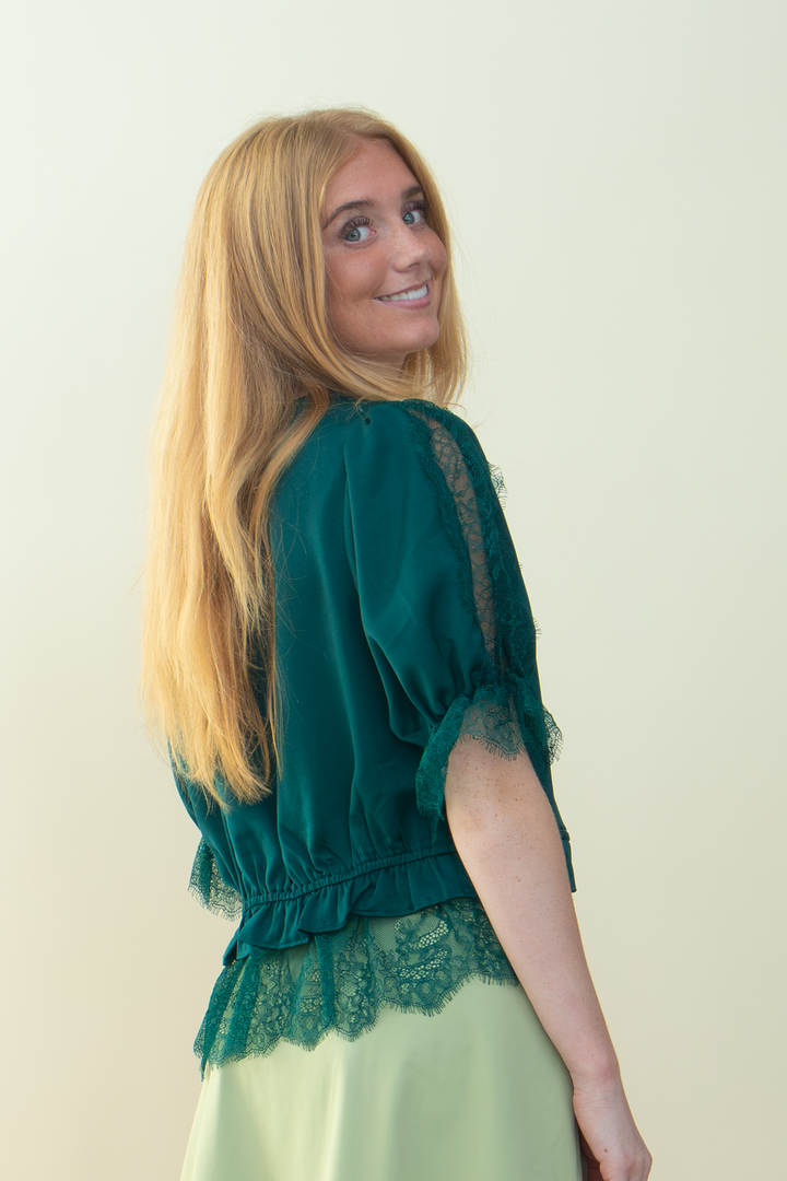 A woman with long blonde hair is shown from the back wearing a dark green lace-trimmed blouse and a pale green skirt, set against a plain light beige background.