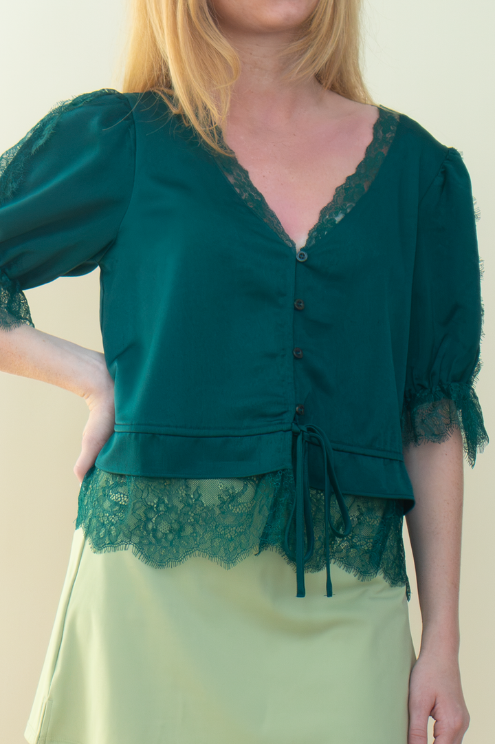 A woman wearing a dark forest green blouse with lace trim, puff sleeves, and a drawstring waist, paired with a light sage green skirt, against a plain beige background.