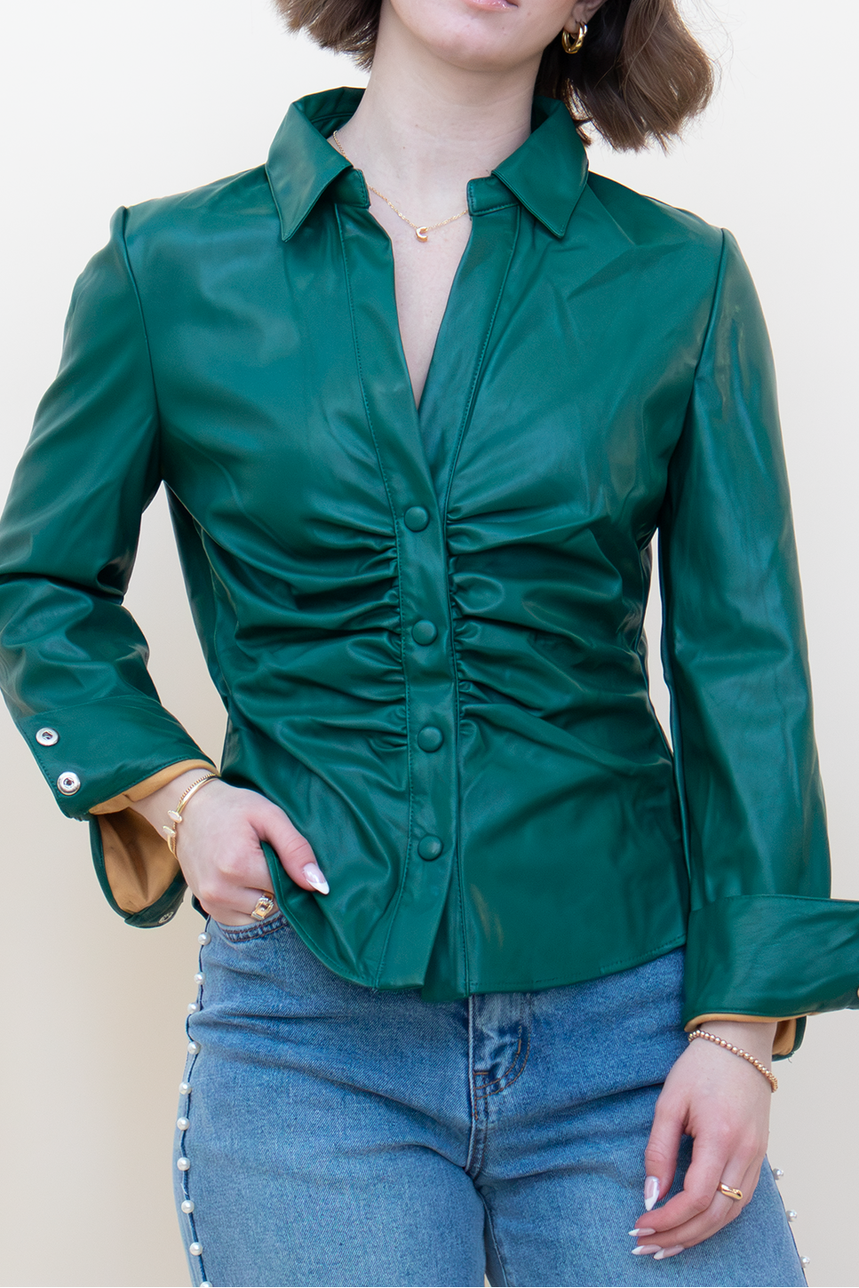 A woman wearing a dark green leather-look button-up shirt with ruching and gold-tone buttons, paired with blue jeans, standing against a plain off-white background.