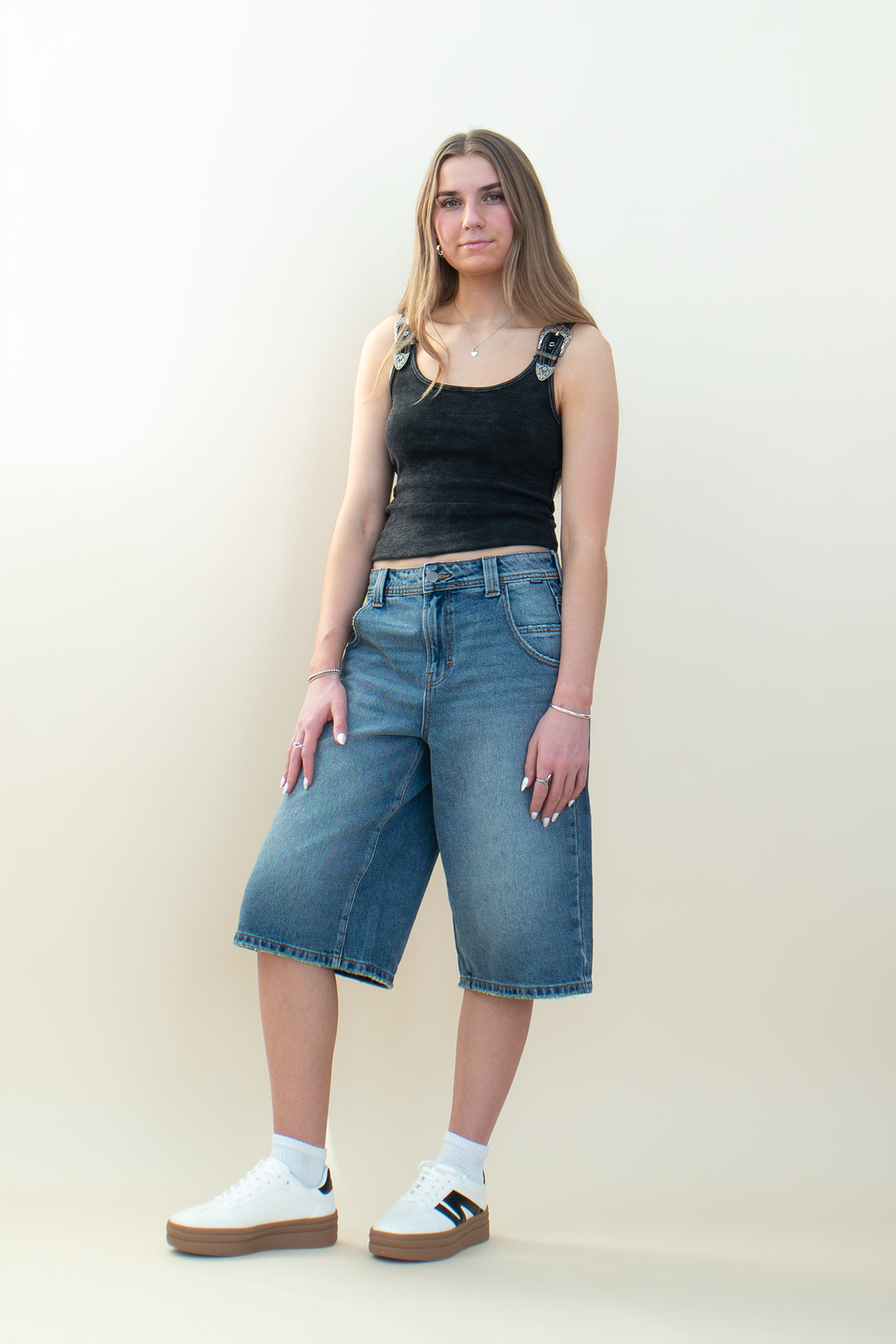 Model wearing medium-wash denim Bermuda shorts with a relaxed knee-length fit, styled with a fitted black tank and sneakers.