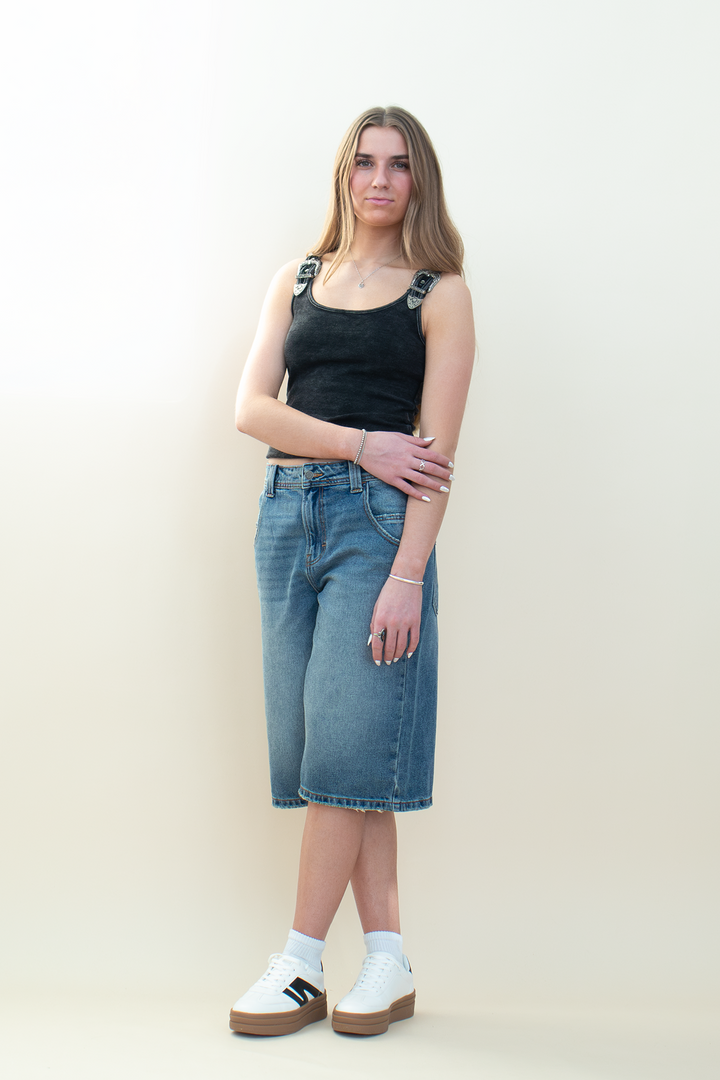 Front view of medium-wash denim Bermuda shorts with a relaxed longline silhouette, paired with a fitted black tank and sneakers.