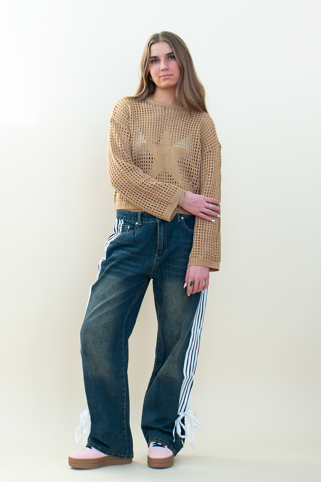 Model wearing a tan open-knit cropped sweater with a large star design, styled with dark wash wide-leg jeans featuring white side stripes.