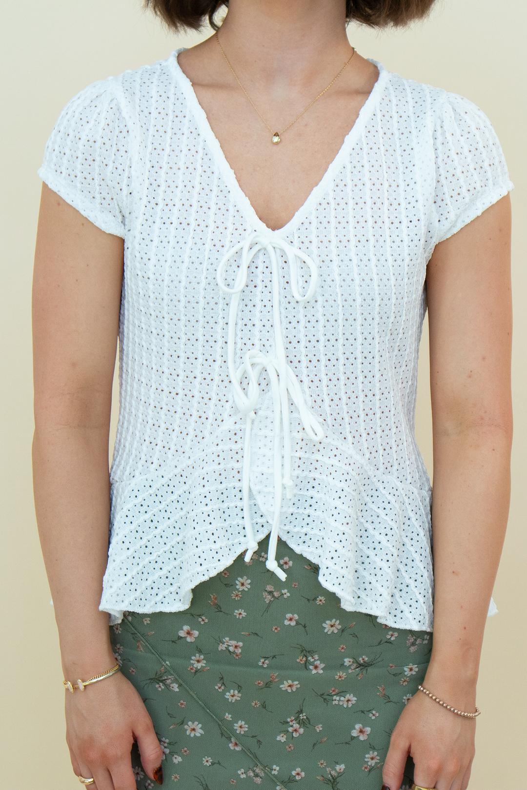 A woman wearing a white short-sleeve crochet knit top with a V-neck and front tie closure, paired with an olive green floral skirt, against a plain beige background.