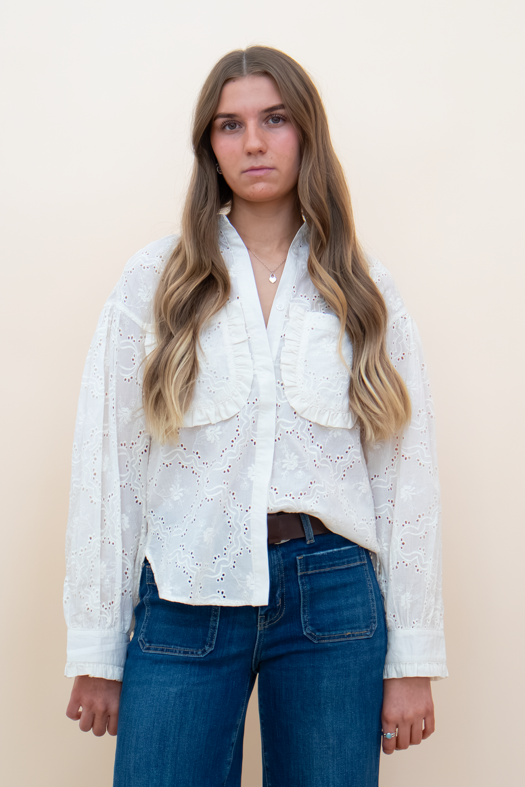 A woman with long wavy blonde hair wearing a white eyelet lace long-sleeve blouse and blue jeans, standing against a plain beige studio background.