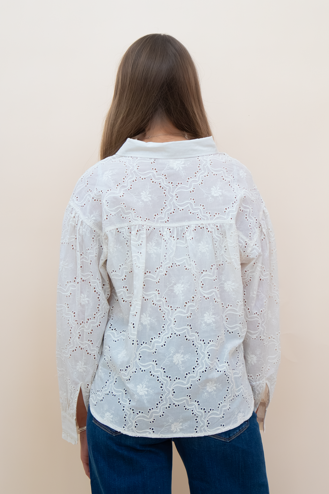 A woman viewed from behind wearing a white long-sleeve eyelet lace blouse with a pointed collar and blue jeans, standing against a plain beige background.