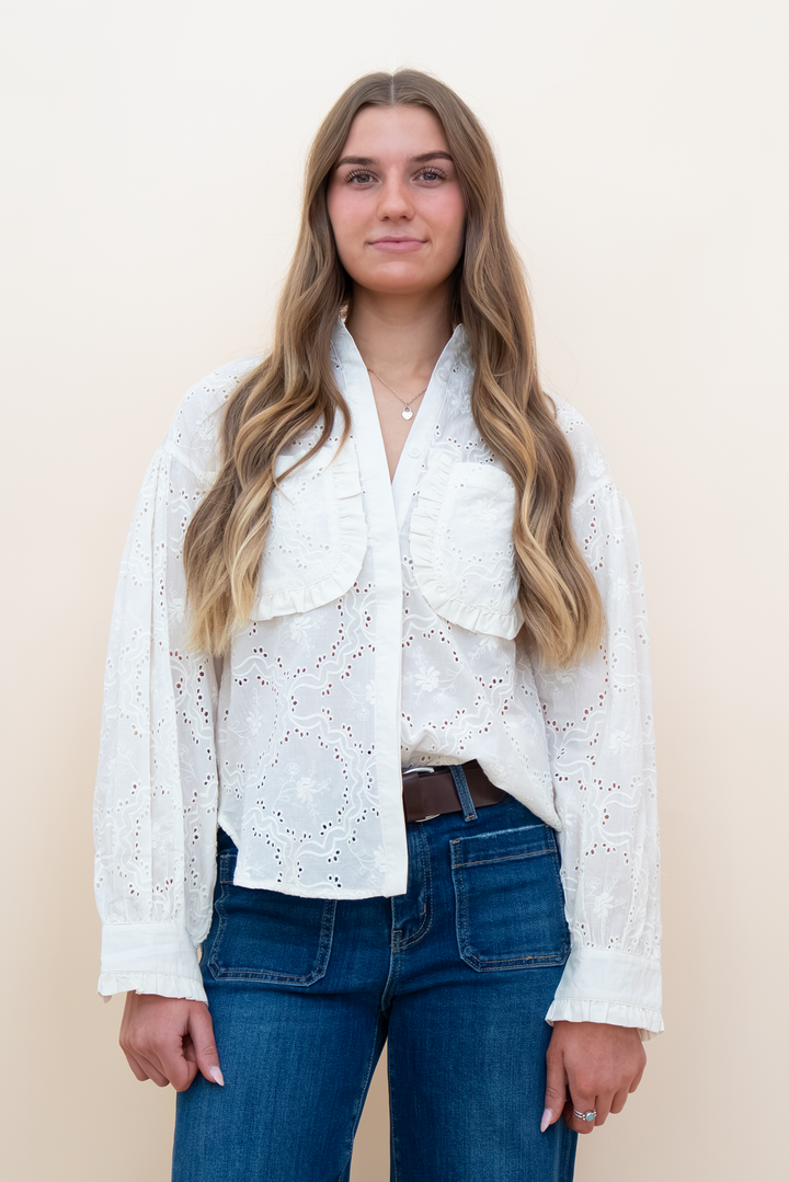 A woman with long wavy blonde hair wearing a white eyelet lace blouse with ruffled details and blue jeans, standing against a plain beige background.