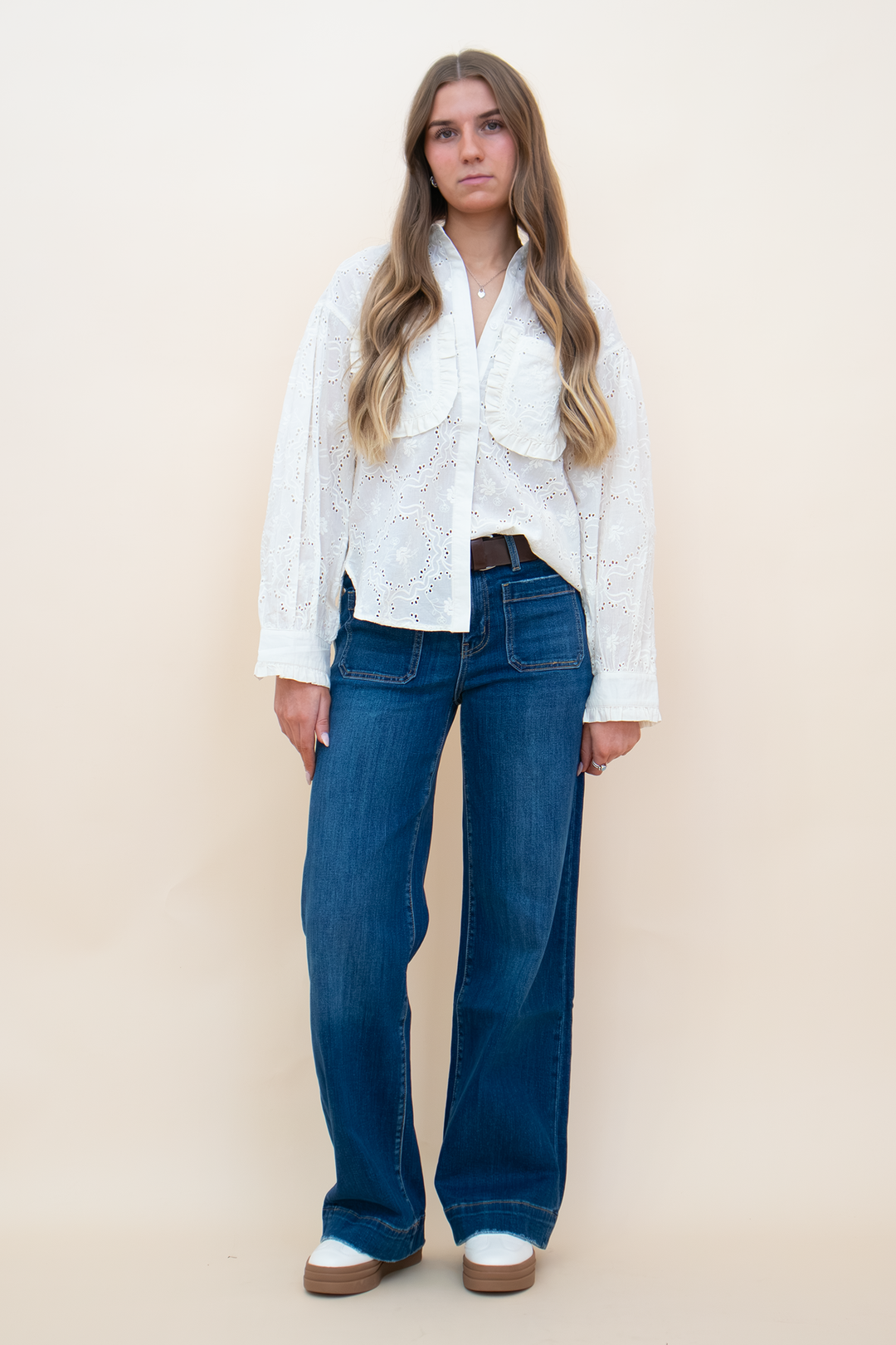 A woman modeling a white long-sleeve lace blouse tucked into blue wide-leg jeans with a brown belt, standing against a solid beige studio background.