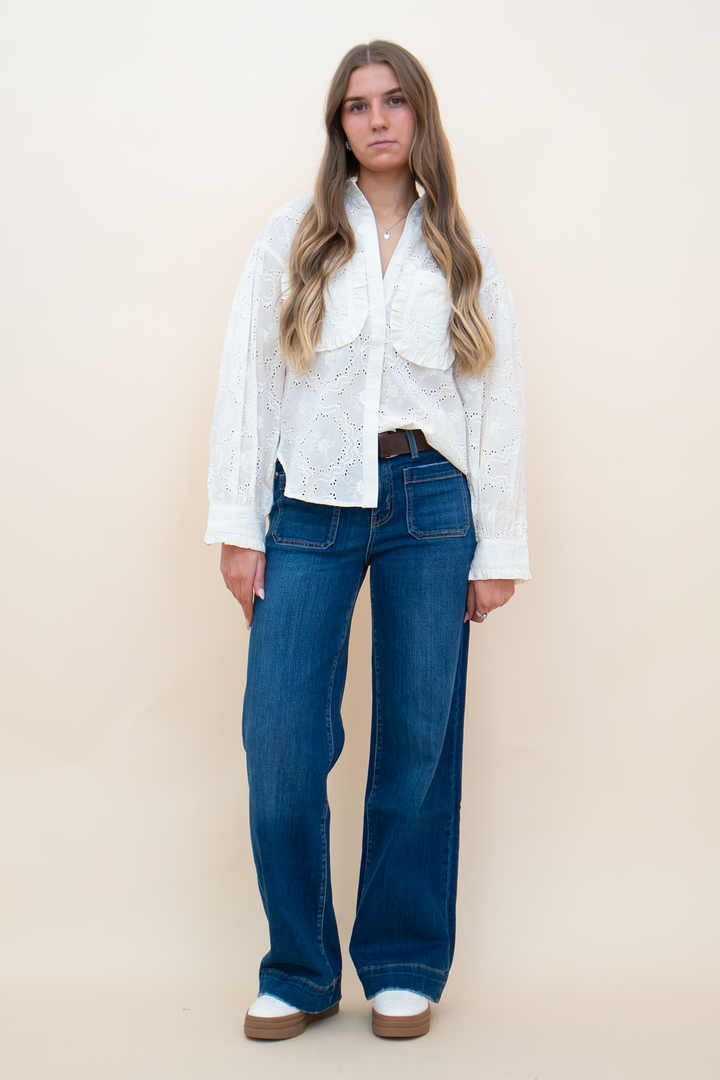 A woman modeling a white long-sleeve lace blouse tucked into blue wide-leg jeans with a brown belt, standing against a solid beige studio background.
