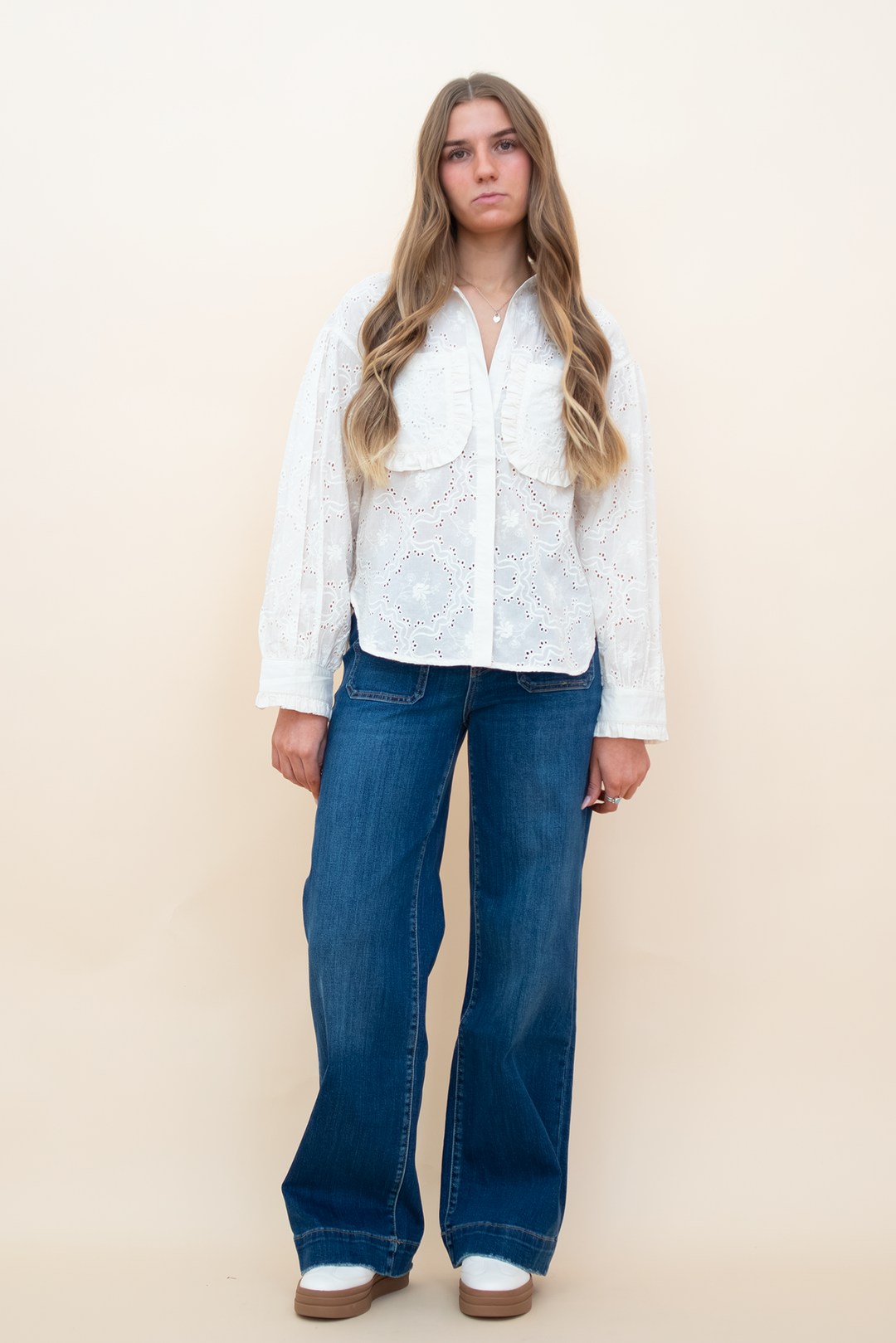 A woman modeling a white long-sleeve lace blouse with button details and wide-leg blue jeans, standing against a solid beige studio background.