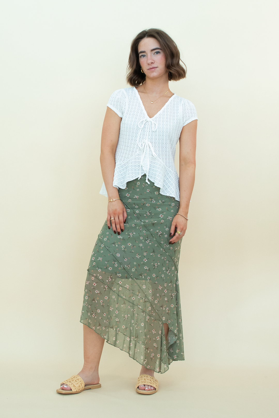 A woman modeling a white short-sleeve tie-front blouse and a green floral midi skirt with a sheer lower overlay, standing against a plain cream studio background.