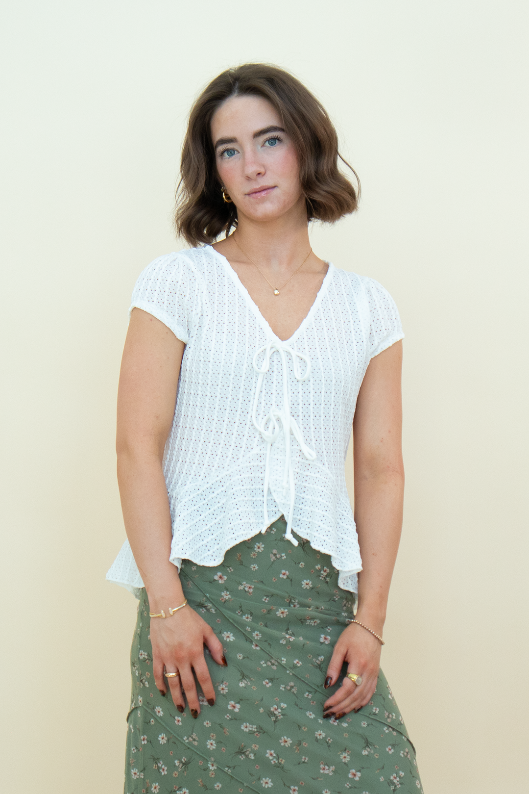 A woman modeling a white short-sleeve crochet knit top with a tie-front detail, paired with an olive green floral skirt, against a plain cream background.