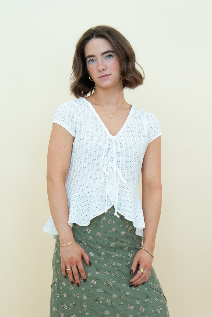 A woman modeling a white short-sleeve crochet knit top with a tie-front detail, paired with an olive green floral skirt, against a plain cream background.