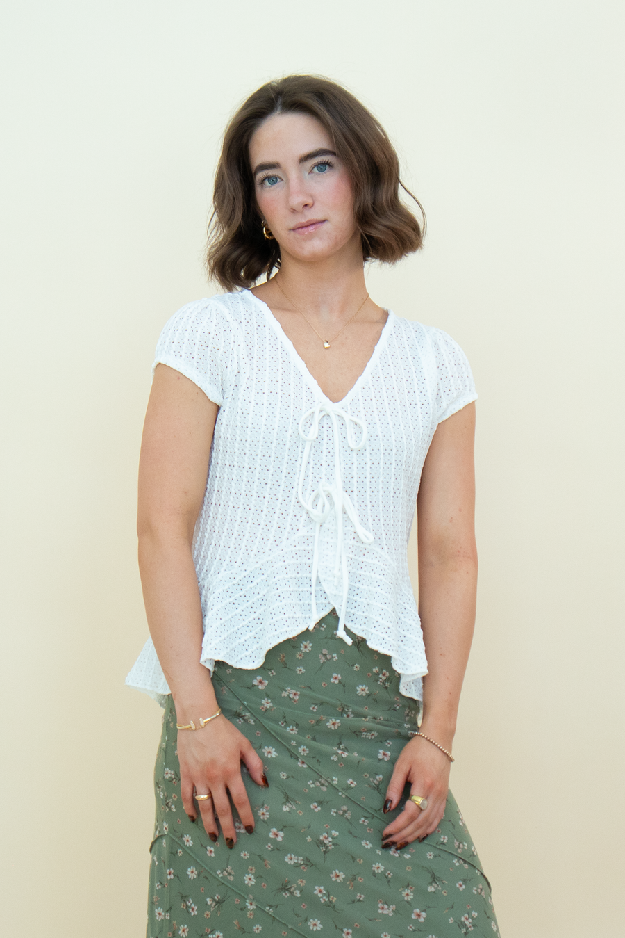 A woman modeling a white short-sleeve crochet knit top with a tie-front detail, paired with an olive green floral skirt, against a plain cream background.